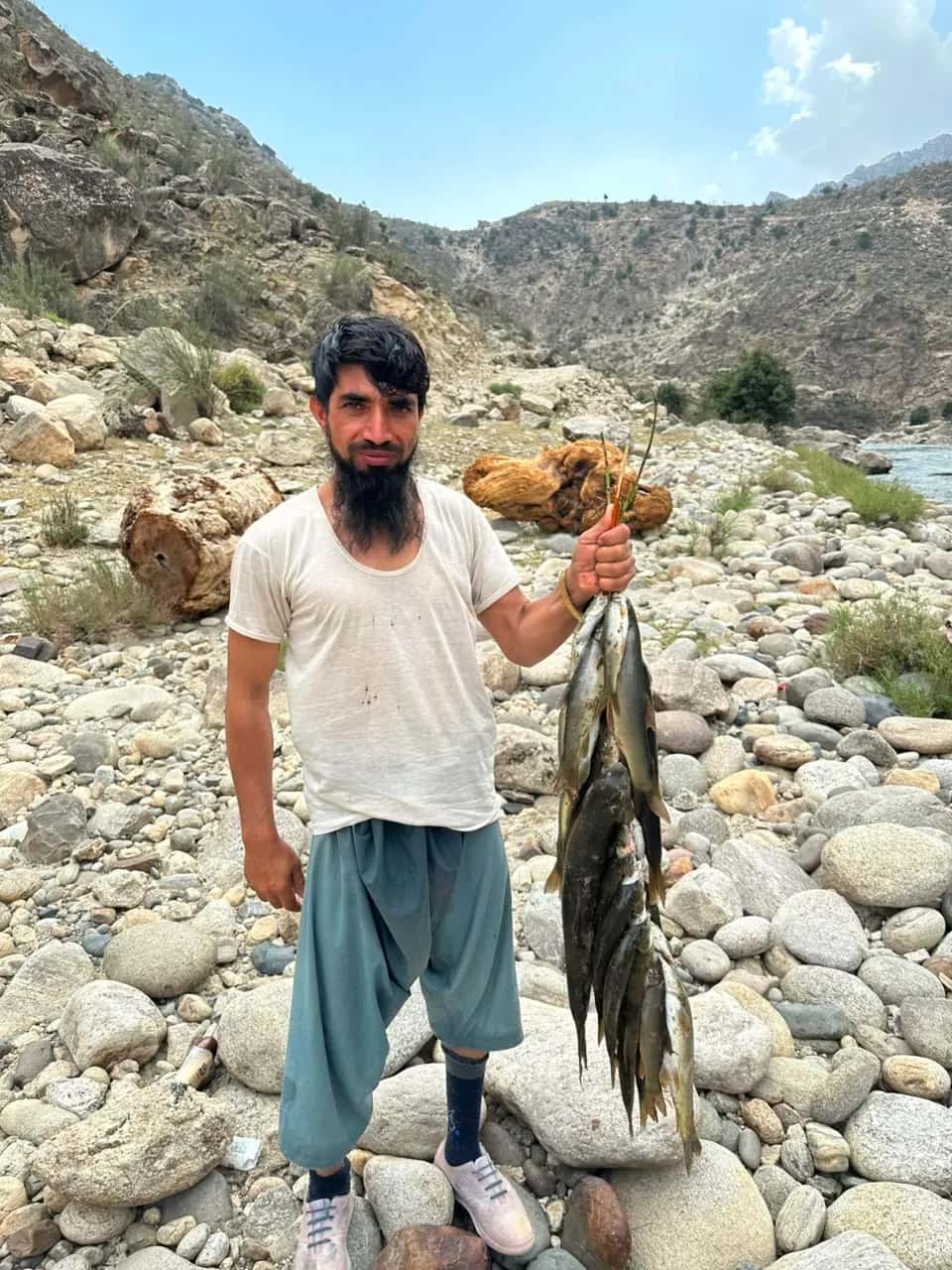A fisherman on a stony riverbank in Mandol holding up a string of fish