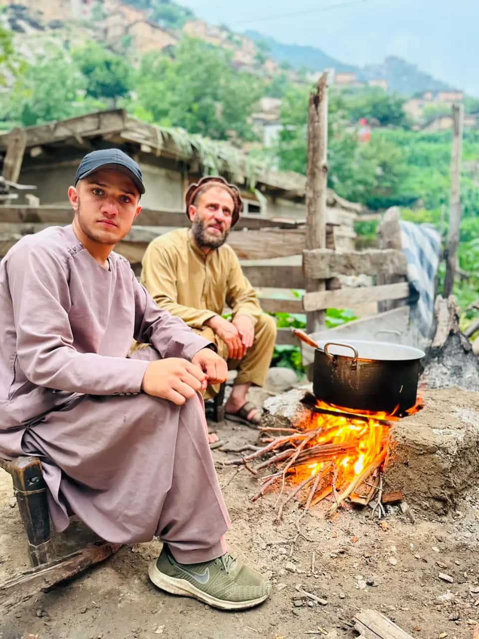Two men beside a blackened pot over an open wood fire, village slopes behind