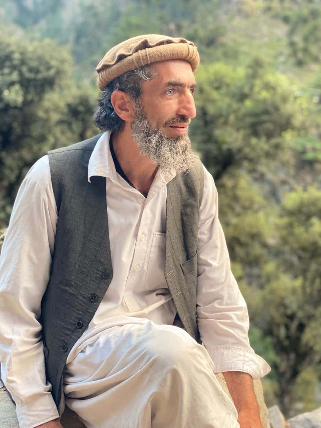 A grey-bearded Nuristani man in a tan pakol and grey waistcoat, forest behind him