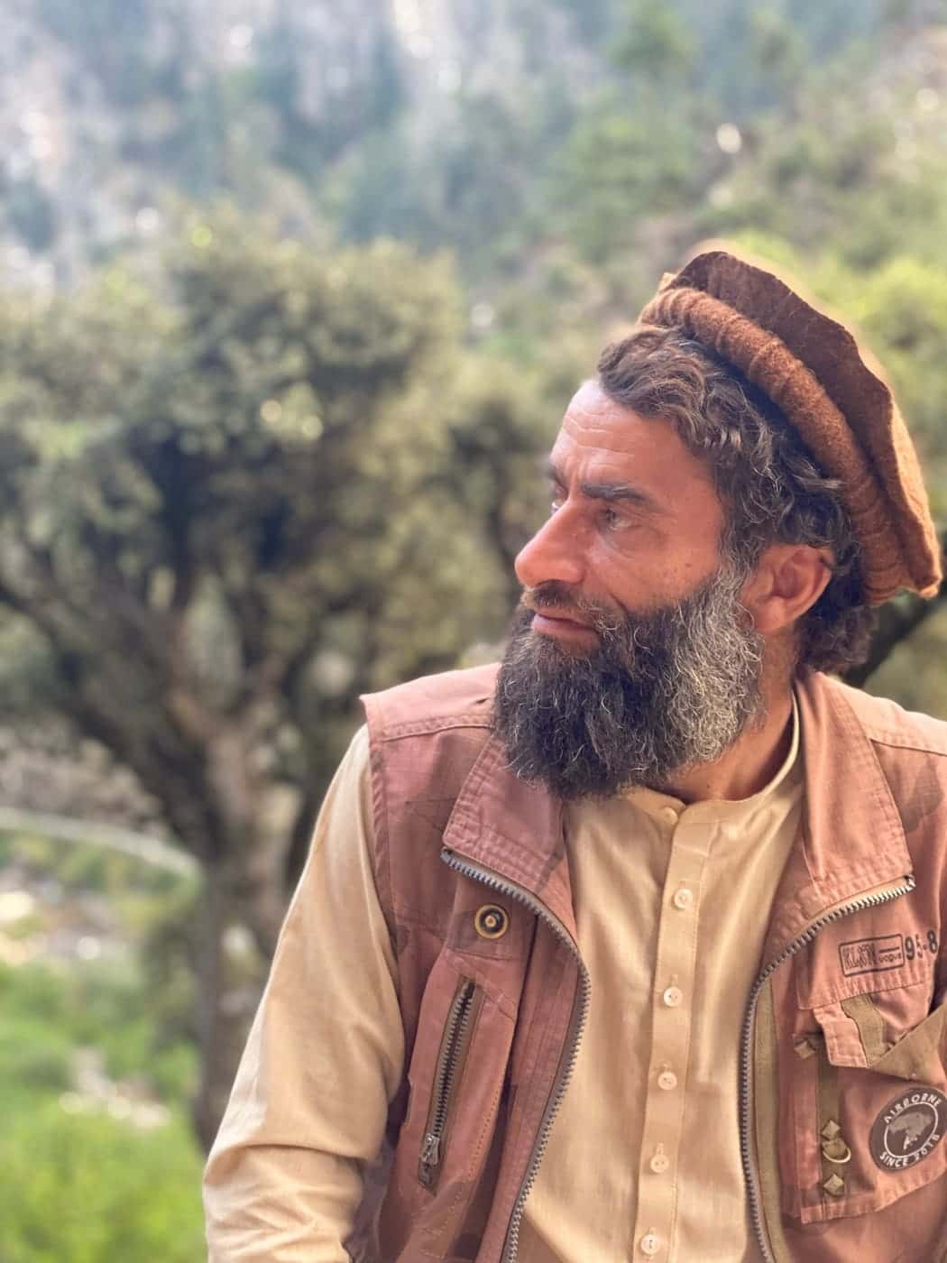 A bearded Nuristani man in a brown pakol rolled high, a cream kamiz and a working vest