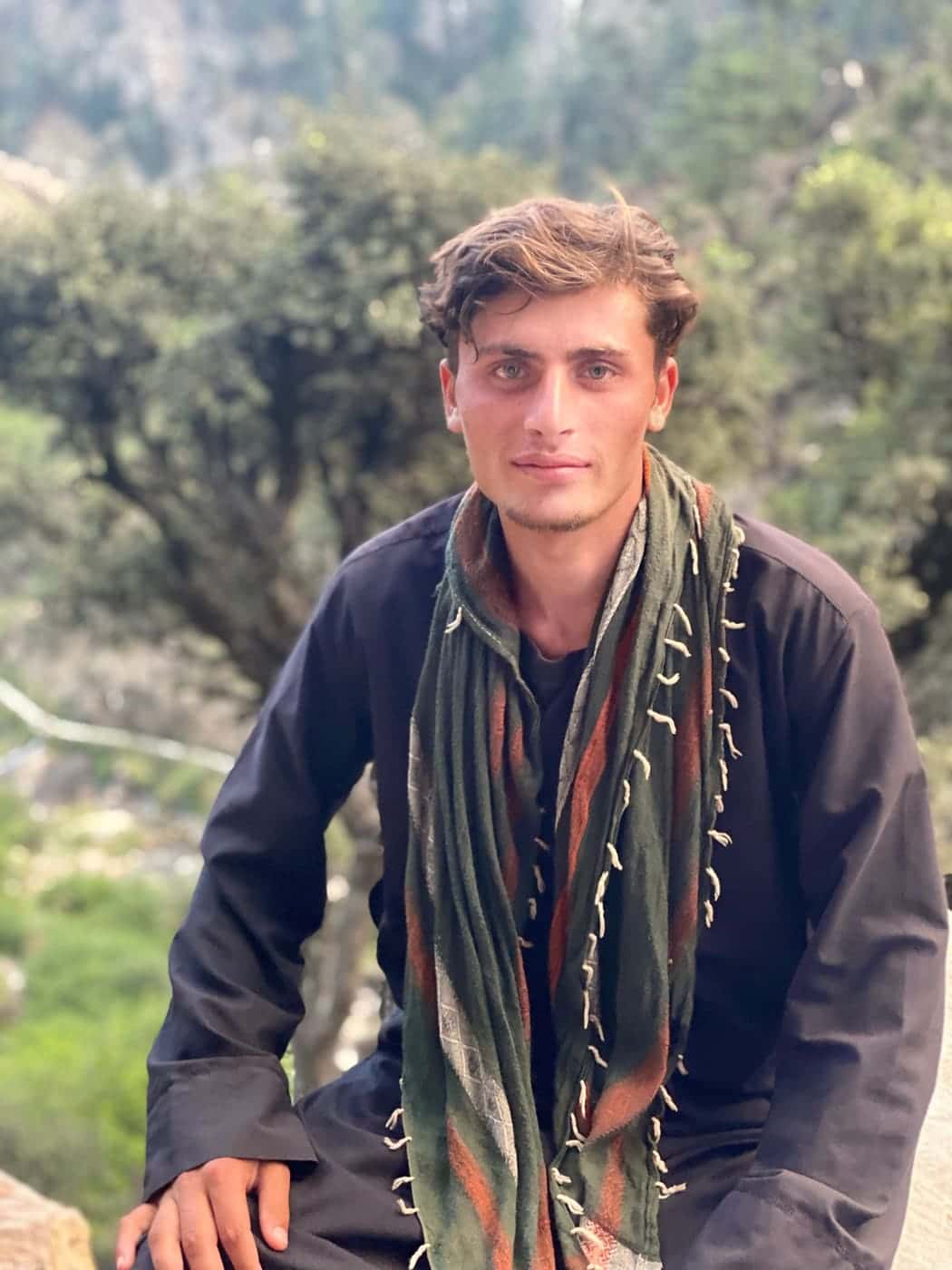 A young Nuristani man in a black kamiz and a green and rust woven wool scarf with knotted tassels
