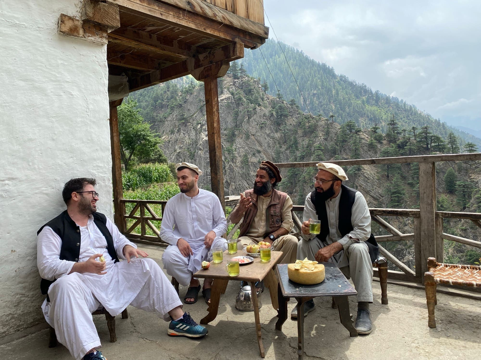 Visitors sharing tea with Nuristani hosts on a timber terrace above a forested valley