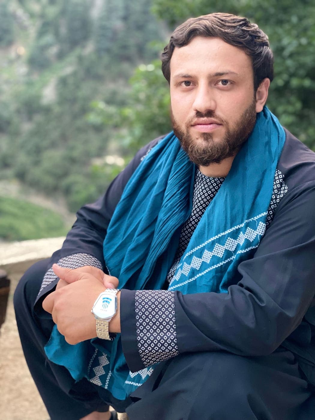 A young man in a black tunic with white embroidery and a blue embroidered scarf