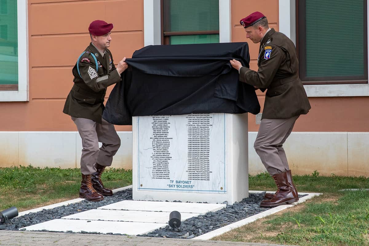 Two soldiers in maroon berets lifting a black cloth from a white marble memorial stone engraved with rows of names, in front of a terracotta building