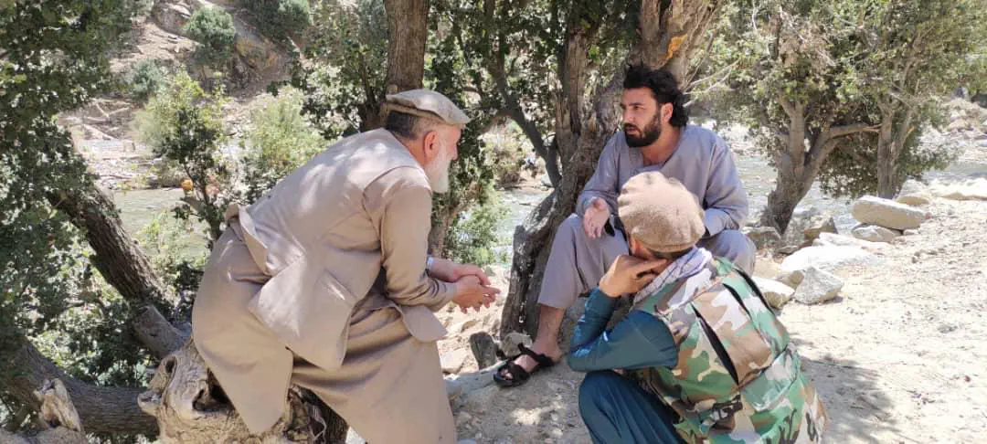 Abdulrahman Chonish sitting under a tree by the river in Mandol, talking with a white-bearded elder in a pakol while another man listens