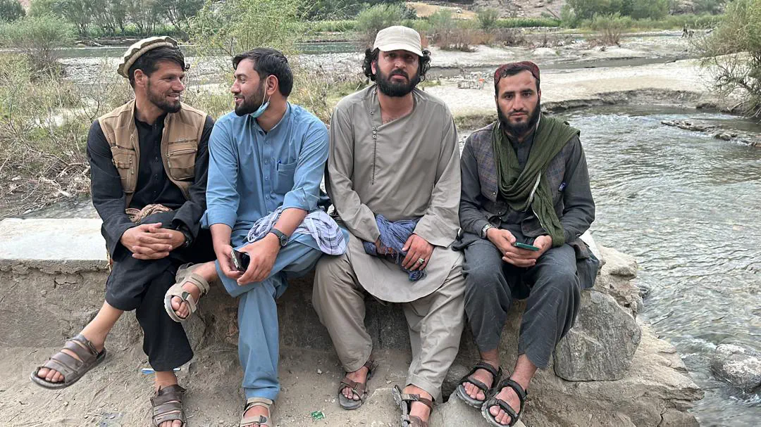 Abdulrahman Chonish sitting on a stone wall by the river in Mandol with three local friends, the river and its gravel banks behind
