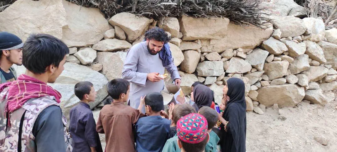 Abdulrahman Chonish handing out gifts to a crowd of village children in the Kuraj valley of Mandol, boys and girls gathered round him in front of a dry-stone wall
