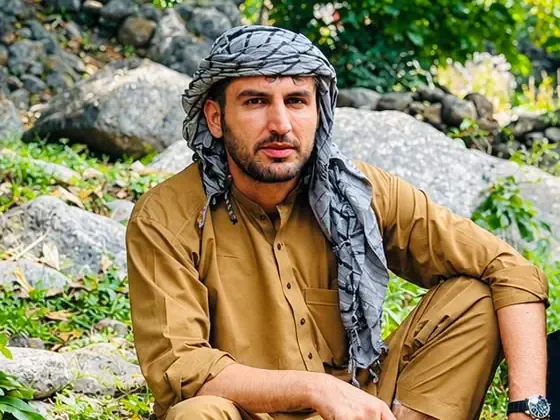 عبدالرحمن چونش، مسئول سفرها و راهنمایی در نورستان، با پیراهن و تنبان خردلی و دستمال چهارخانهٔ خاکستری، بر سنگی در درهٔ پشاگر ولسوالی نورگرام نشسته است