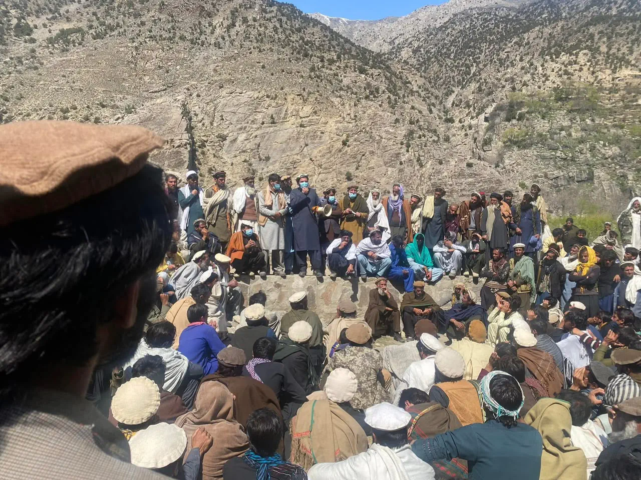 جرگهٔ بزرگی در مندول: مردان قوم نشسته بر زمین و ایستاده در امتداد دیوار سنگی زیر کوهی صخرهای، و سخنرانی با بلندگو در میان