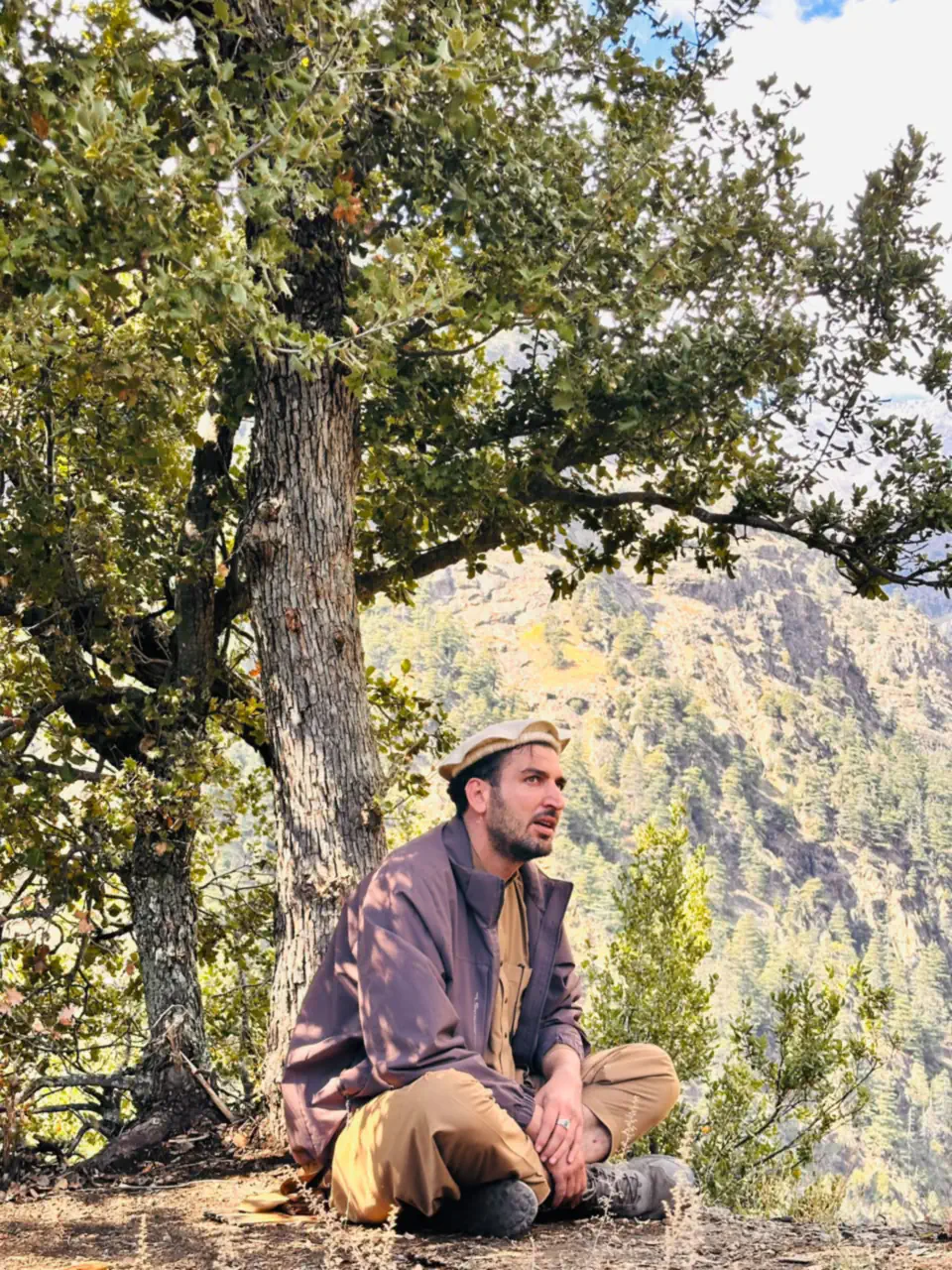 Abdulrahman Chonish in a cream pakol and purple jacket sitting cross-legged under an oak tree high in the Sewketra valley of Waygal, cedar-forested slopes behind