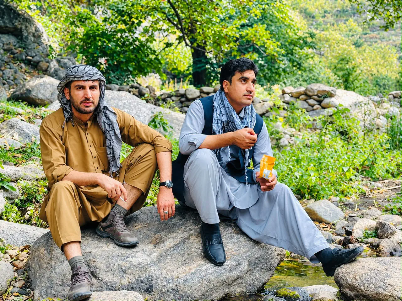 Abdulrahman Chonish and a friend sitting on a boulder beside a stream in the Pashagar valley of Nurgaram, green trees and a dry-stone wall behind them