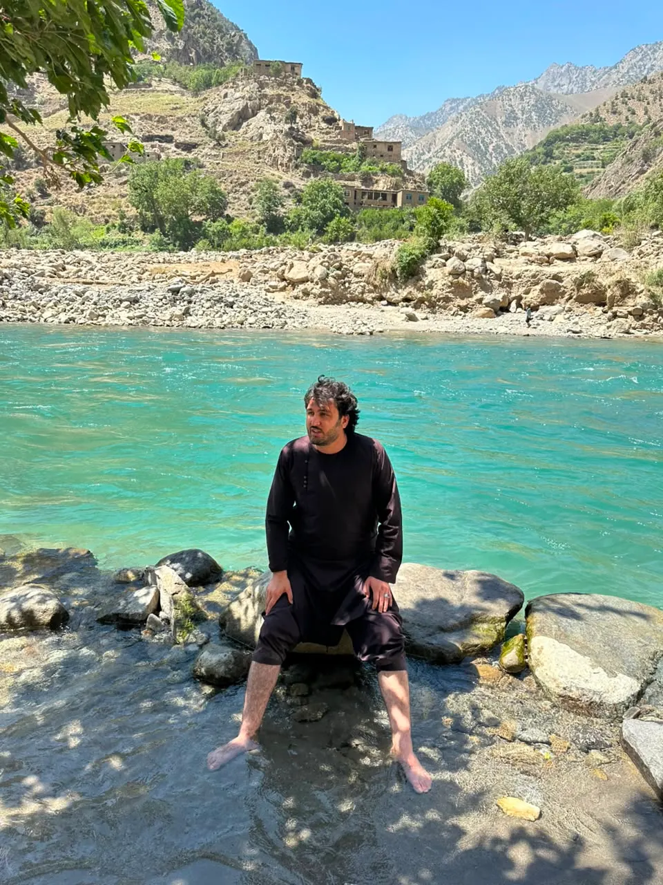 Abdulrahman Chonish sitting on rocks with his feet in a turquoise river in the Kuraj valley of Mandol, a hillside village behind