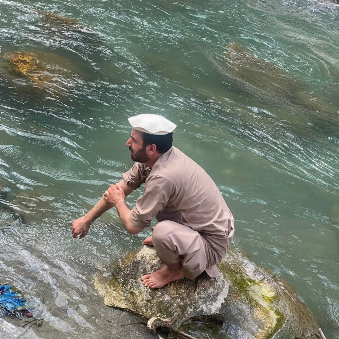 Abdulrahman Chonish in a white pakol crouched barefoot on a rock in a clear green river in Mandol, washing his arms for wudu before prayer