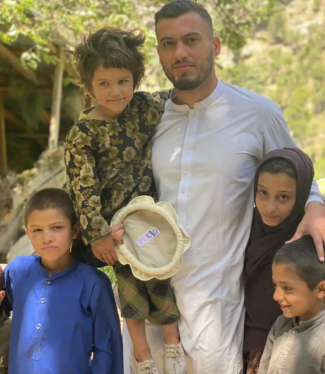 Ahmed Bilal, in a white shalwar kameez, with five children of the Sewketra valley in Waygal district: a small child in a floral kameez holding a new pakol, a girl in a brown headscarf under his arm and three boys, a timber house and the forested valley side behind