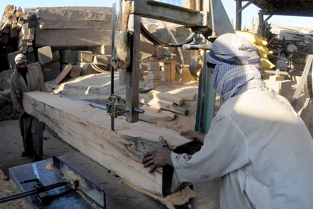 Two men guiding a large cedar beam through a band saw at a timber yard in Asadabad, stacked logs behind them