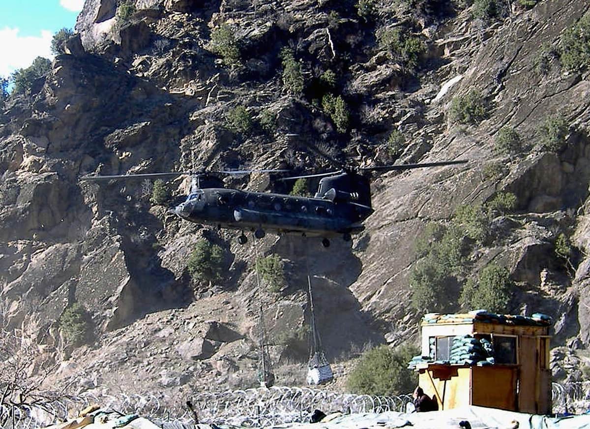 A twin-rotor Chinook helicopter coming in to land beneath a sheer rock face, a sandbagged wooden guard post and coiled wire in the foreground