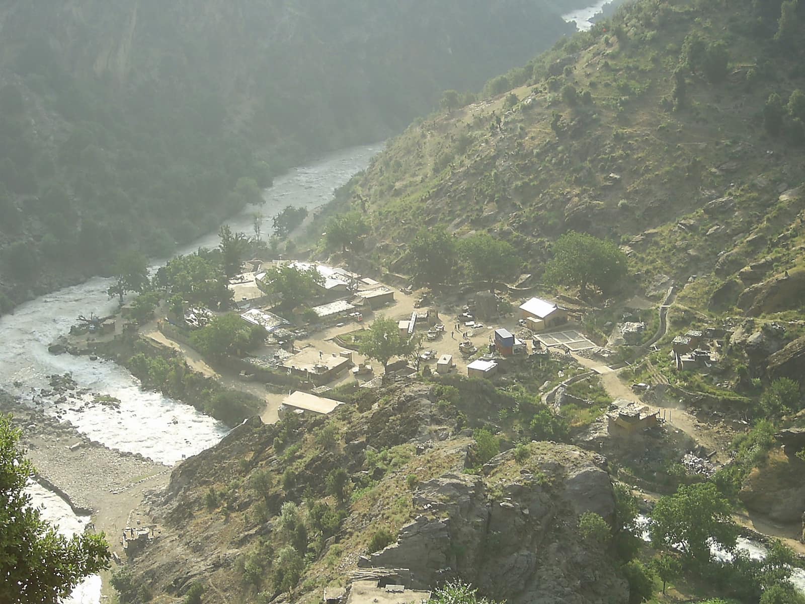 Combat Outpost Keating seen from the mountain above: a cluster of small buildings and trees on a bend of the Landai Sin river at the bottom of a steep green valley