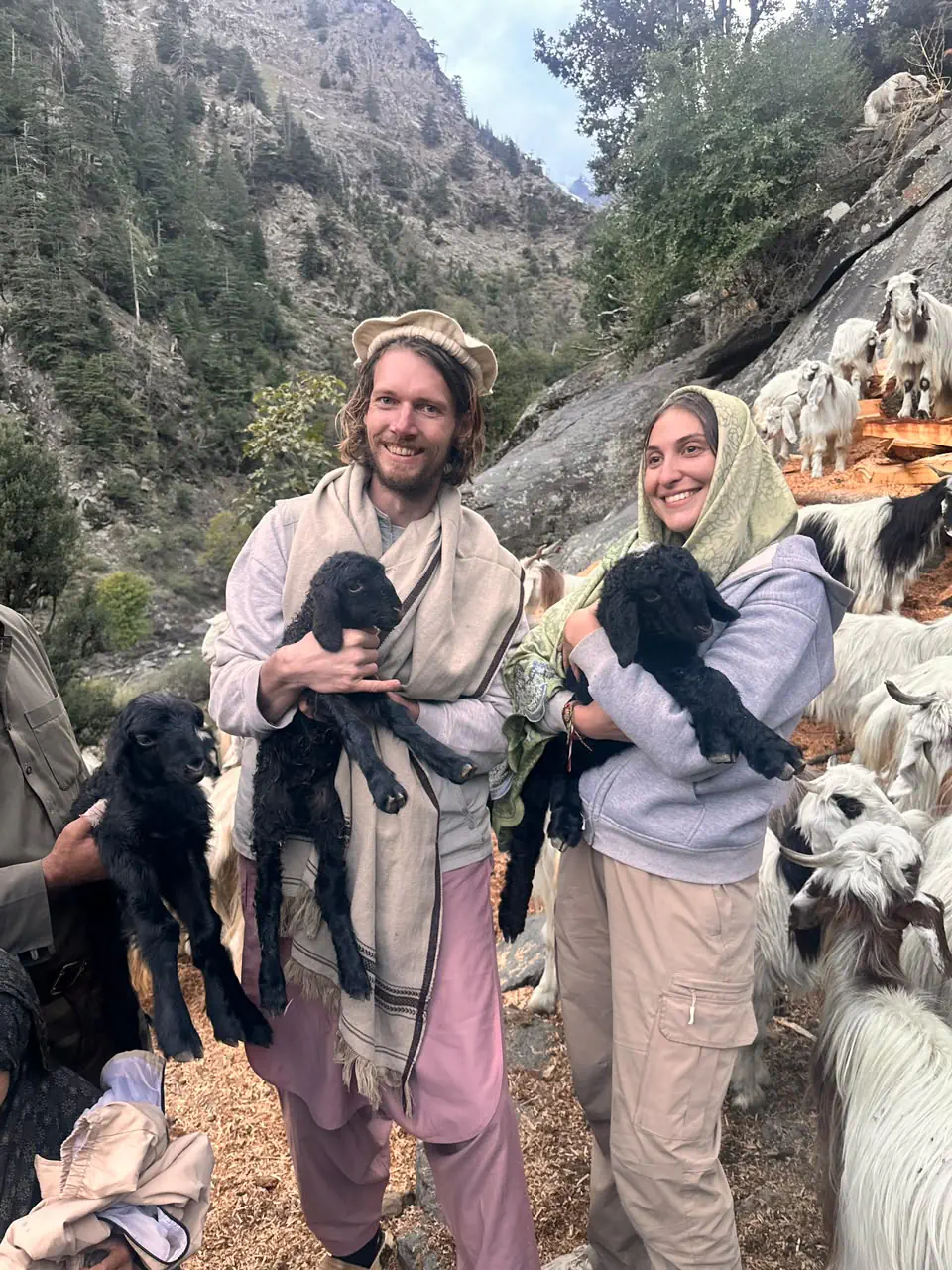 A smiling couple from the Netherlands, he in a pakol and shawl, she in a headscarf, each holding a black newborn kid among long-haired goats in the Sewketra valley of Waygal
