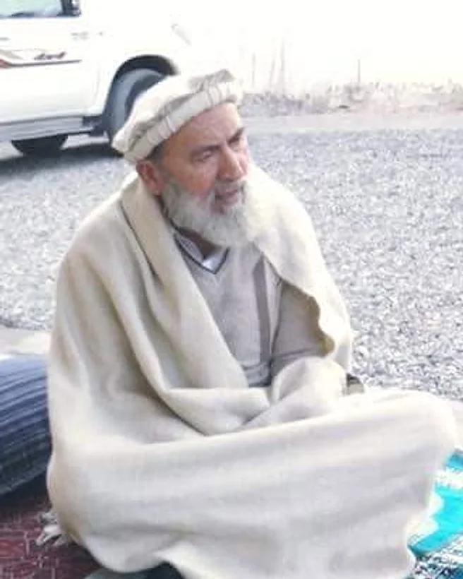 مرحوم انجنیر امیرجان نورستانی، جیولوجیست و از بزرگان نورستان از وایگل، با پکول سفید و شال کریمی، چهارزانو بر قالین نشسته است