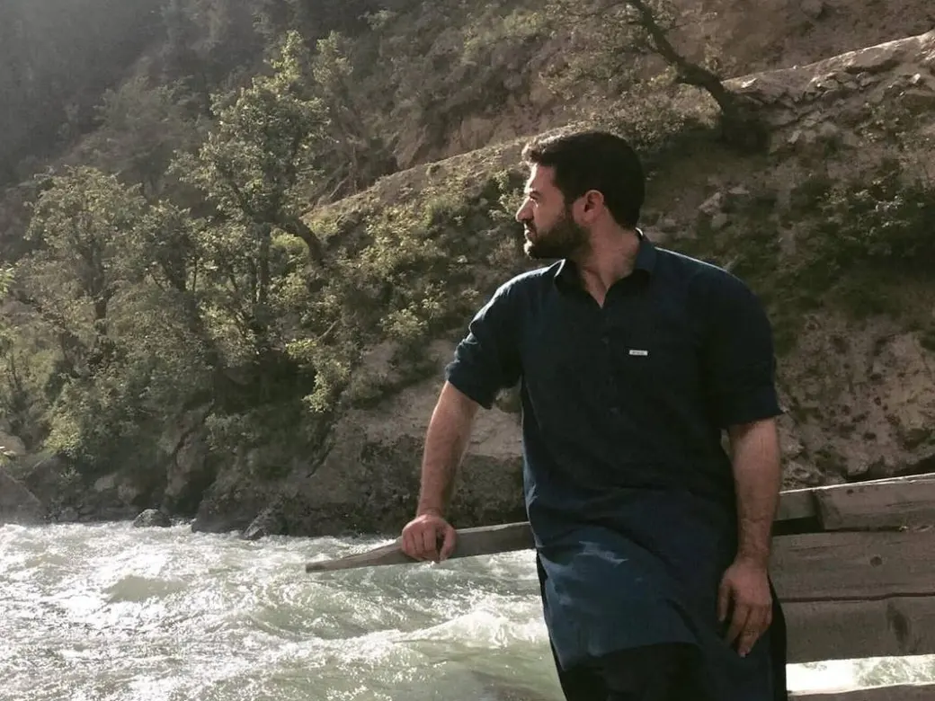 Habib Rahman, Nuristani's business manager for Europe, in a dark blue shalwar kameez, leaning on the rail of a wooden bridge over the white water of the Parun river