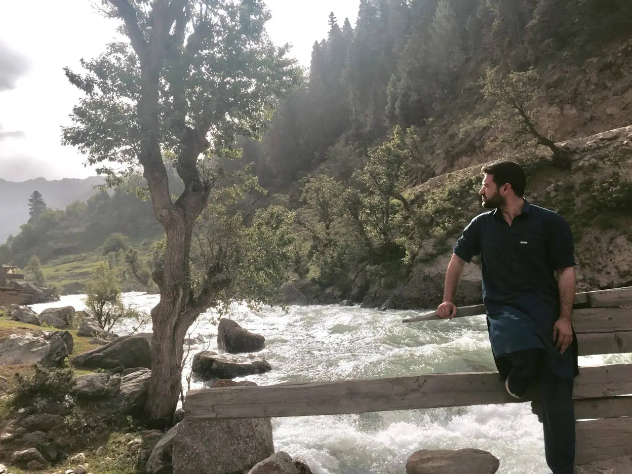 Habib Rahman, in a dark blue shalwar kameez, sitting on the rail of a wooden bridge over the Parun river and looking upstream, white water over boulders, a tree on the bank and cedar forest on the valley side