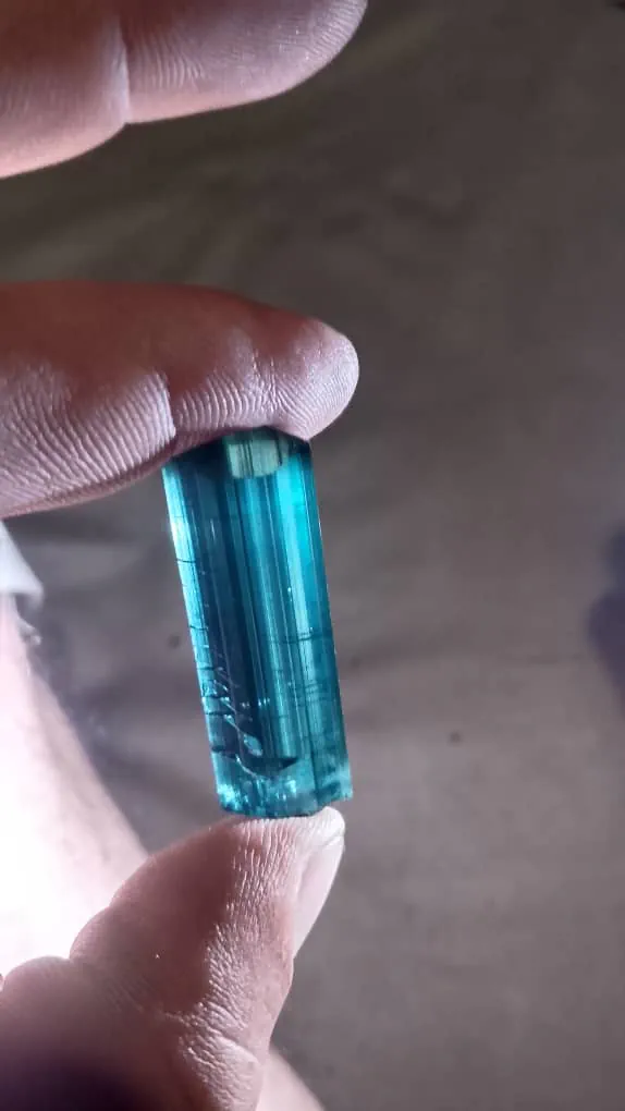 A long blue tourmaline crystal, indicolite, from the Yinshaw valley in Waygal, Nuristan, held between finger and thumb, its faces striated along the length