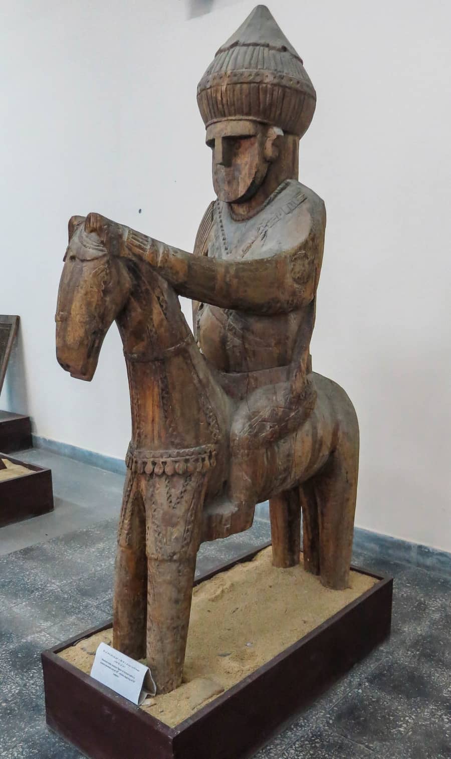 A restored wooden figure of a man in a tall cap seated on a horse, about life size, on a plinth in the National Museum of Afghanistan
