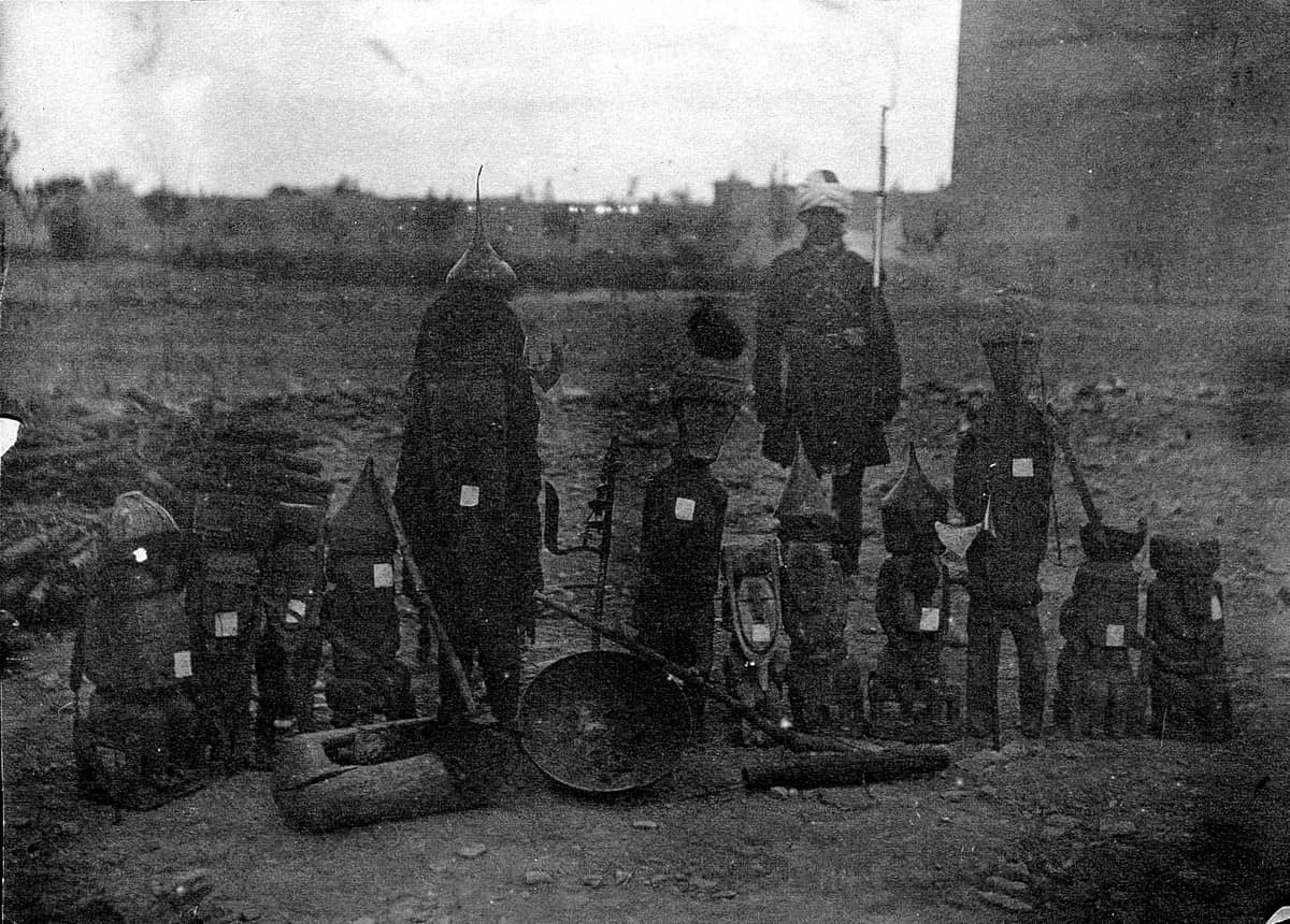 A row of carved wooden figures of different heights standing on open ground with labels attached, a soldier standing behind them, photographed in Kabul in the 1890s