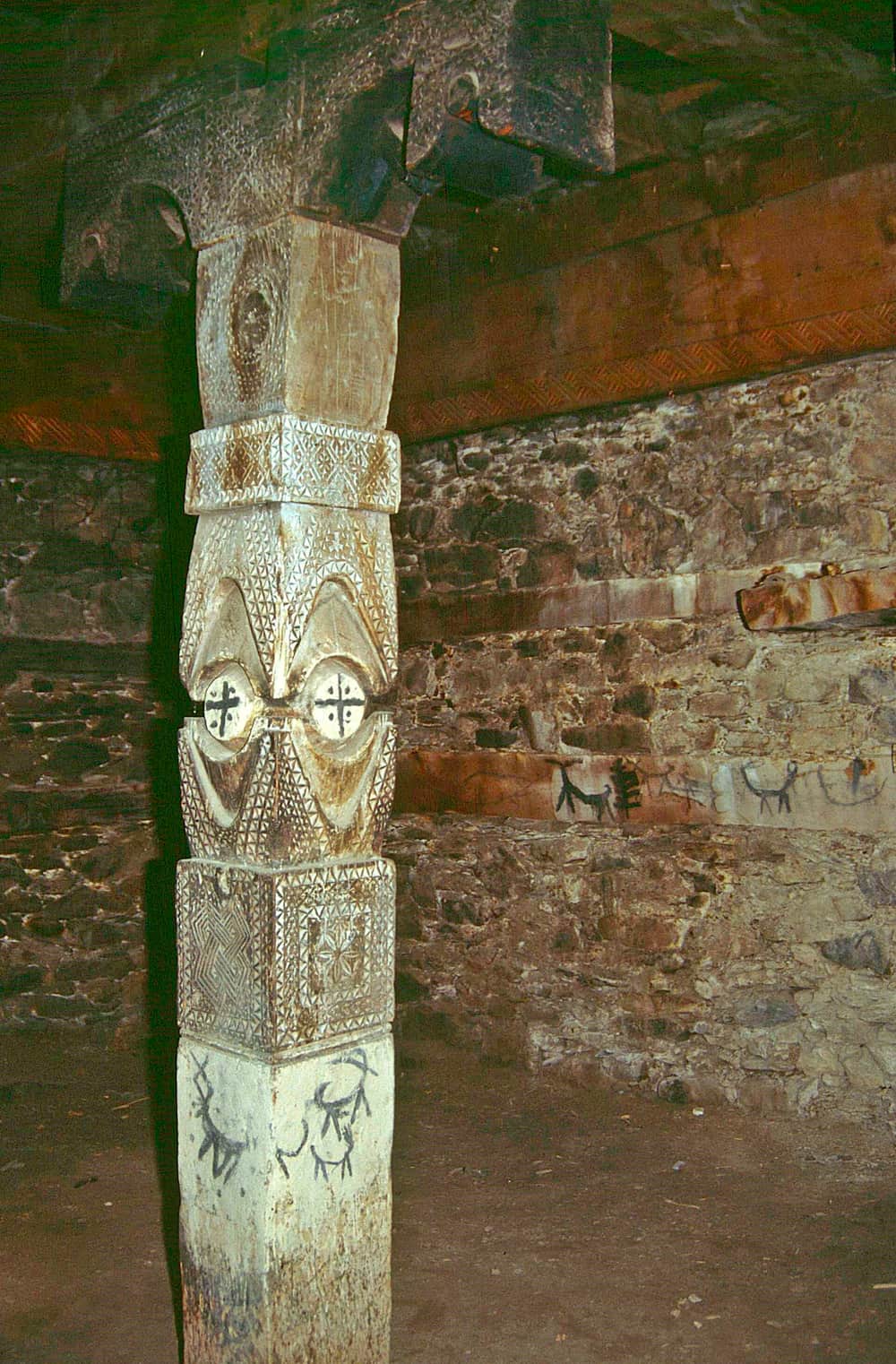 A wooden pillar carved with bands of chip carving and two rosettes inside a stone-walled Kalash dance hall, goats painted on the wall behind it