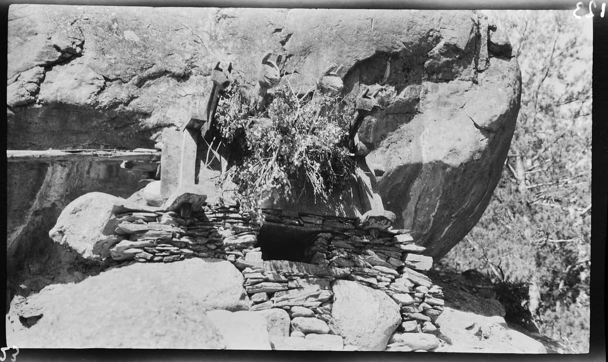 A 1929 photograph of a Kalash altar built of dry stone under a great boulder, with four carved wooden horses' heads and a bundle of branches above the offering hole