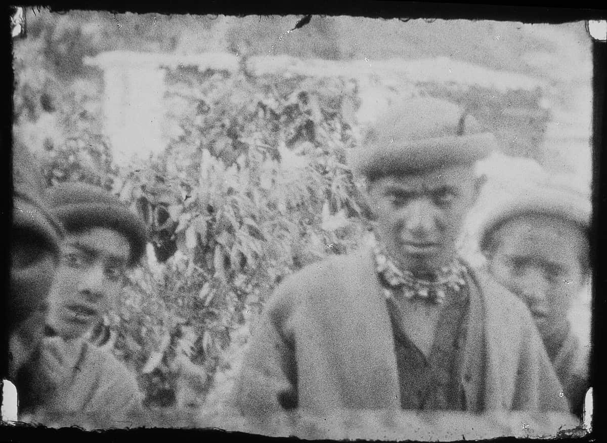 A grainy 1929 photograph of Kalasha youths in caps and bead necklaces standing before an altar dressed with walnut branches