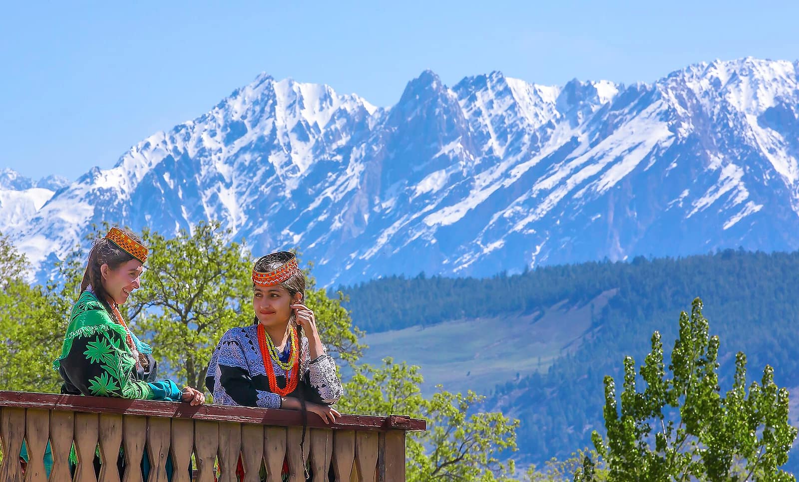 Two Kalash women in embroidered black dresses and beaded headdresses at a wooden rail, with the snow peaks of Chitral behind them