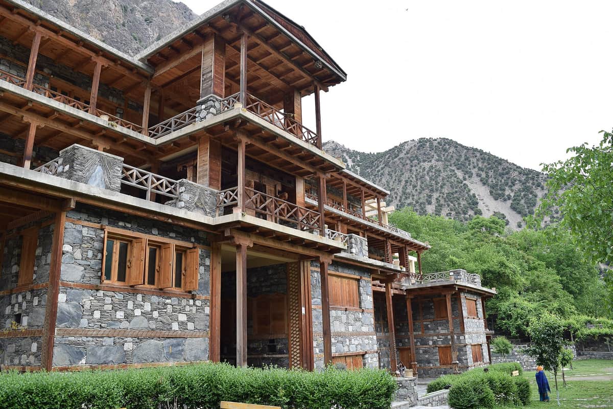 The Kalasha Dur in Bumburet: a large building of grey stone and timber with carved wooden balconies on three floors, trees and a mountain behind