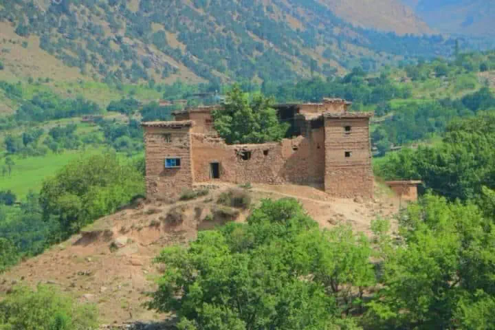 An old stone house with high walls and small windows standing alone on a bare knoll in Kantiwa, green trees and fields around it