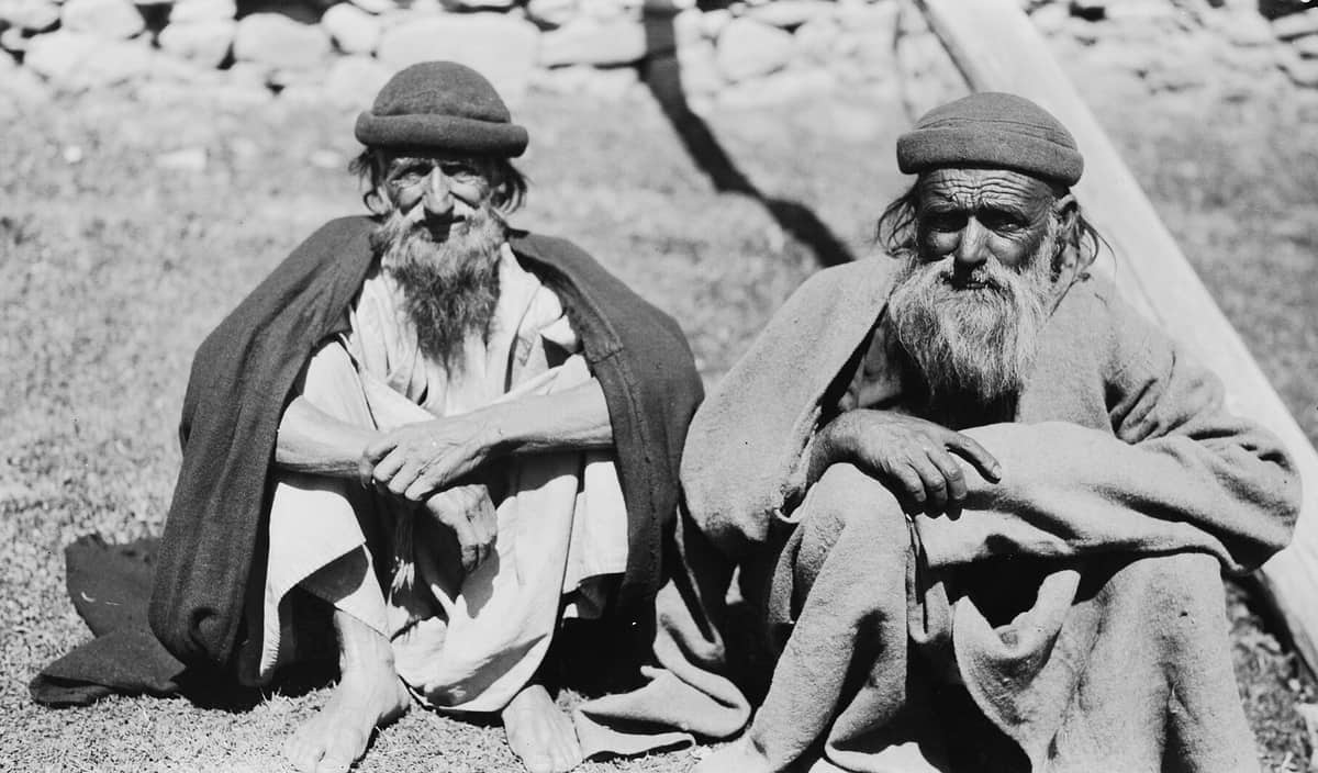 A 1929 photograph of two bearded old men in round wool caps and heavy cloaks sitting side by side on the grass in front of a stone wall