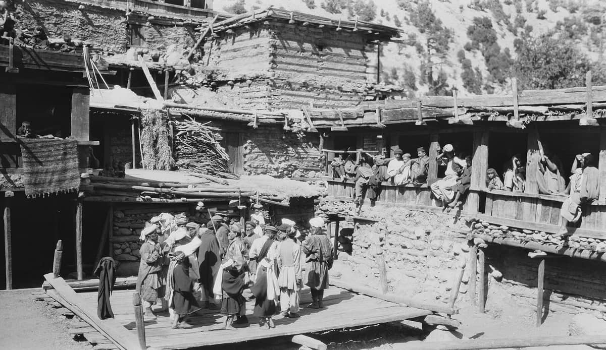 A 1929 photograph of a crowd of men dancing on a raised plank floor between timber and stone houses, some holding axes, with onlookers on the verandas