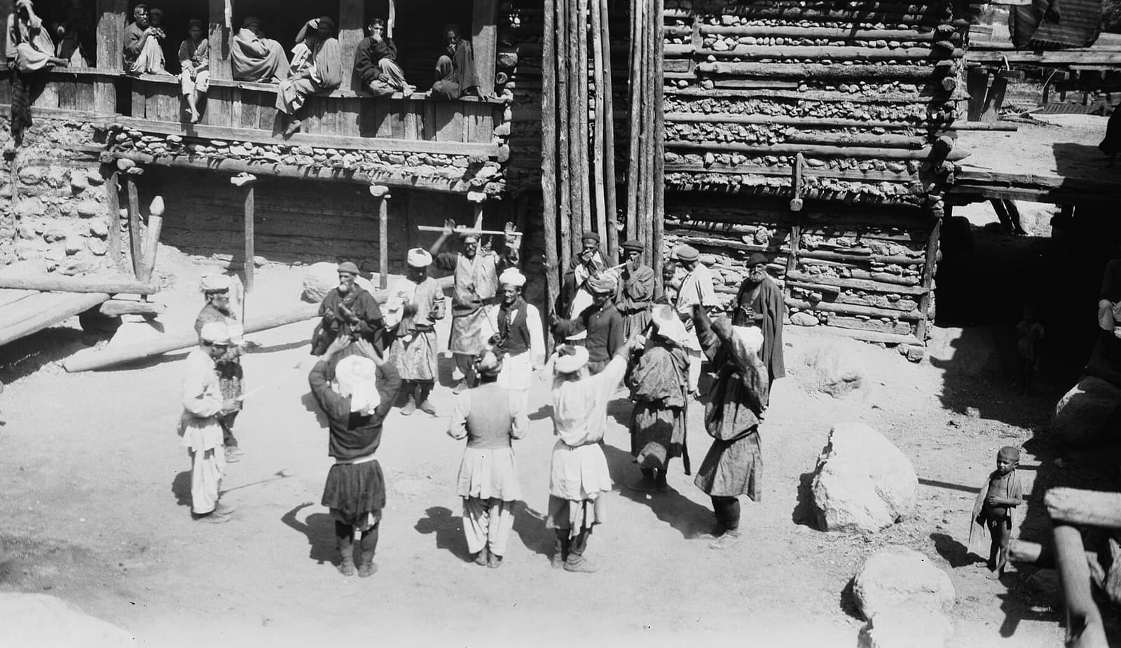 A 1929 photograph of Kati men dancing in a ring in the yard of a timber house, arms raised, with spectators watching from the veranda above