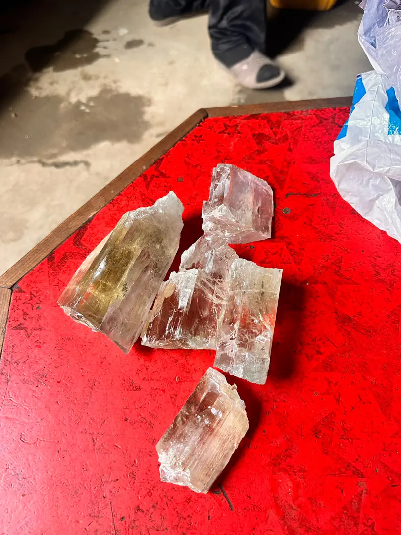 Six rough spodumene crystals from the Mawi mine in Nuristan on a red table: pale pink kunzite, colourless and golden yellow, each grooved along its length with etched, stepped ends