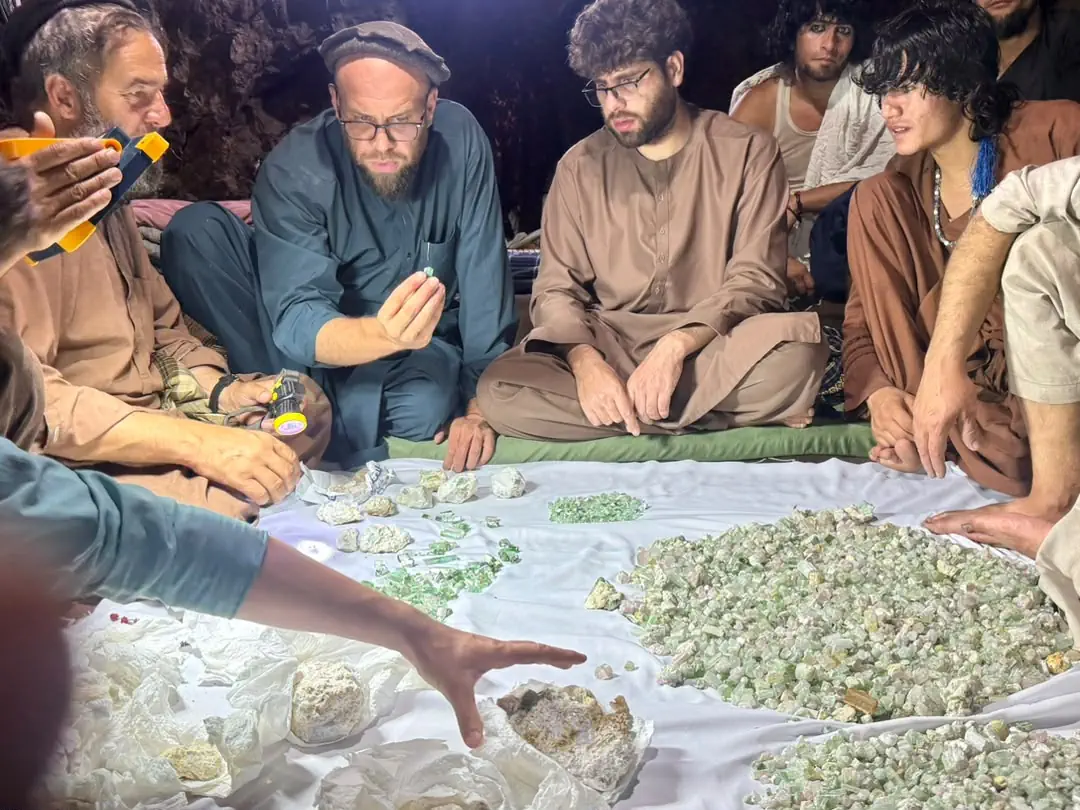 A buyer at the Mawi mine in Du Ab, Nuristan, holding a green tourmaline crystal up to a torch to grade it, with miners and traders seated round the heaps of rough on a white cloth