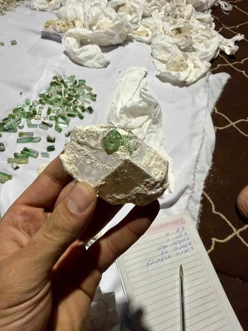 A green tourmaline crystal set in a fist-sized piece of white pegmatite rock from the Mawi mine in Du Ab, Nuristan, held in a hand above a cloth of loose green tourmaline crystals and a notebook of weights