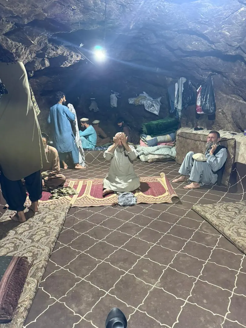 The miners' quarters inside the Mawi mine in Du Ab, Nuristan: a chamber cut into the rock with carpets on the floor, bedding stacked against the wall, a lamp overhead, men resting and one at prayer