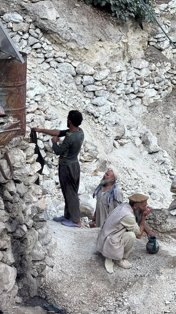 Miners at a camp on the pale pegmatite waste slope of the Mawi mine in Du Ab, Nuristan, one hanging clothes beside a solar panel and an oil drum, two older men resting below the broken rock