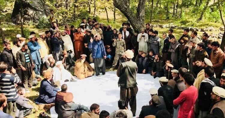 A large circle of men seated and standing at an outdoor village council