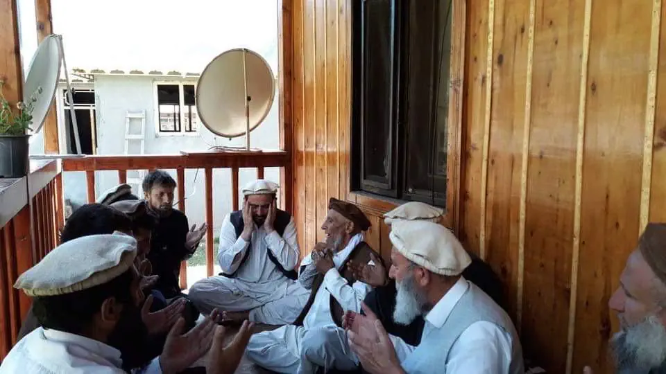 Obaid Nuristani and elders in pakols seated on a timber veranda in Parun, raising their hands in dua, one with his hands over his face