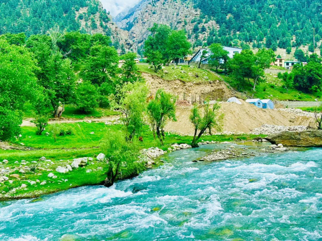 The turquoise Parun river running fast past willows and earth banks, houses among the trees beyond