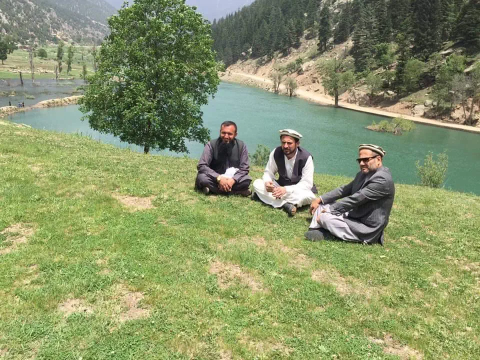 Obaid Nuristani and two friends in pakols and waistcoats sitting on the grass above the turquoise Parun river, pine forest on the slopes beyond