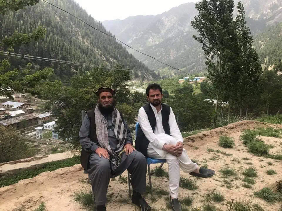 Obaid Nuristani seated beside an elder, a Nuristani uncle, on a hillside above a village in the Parun valley, pine-forested mountains rising behind