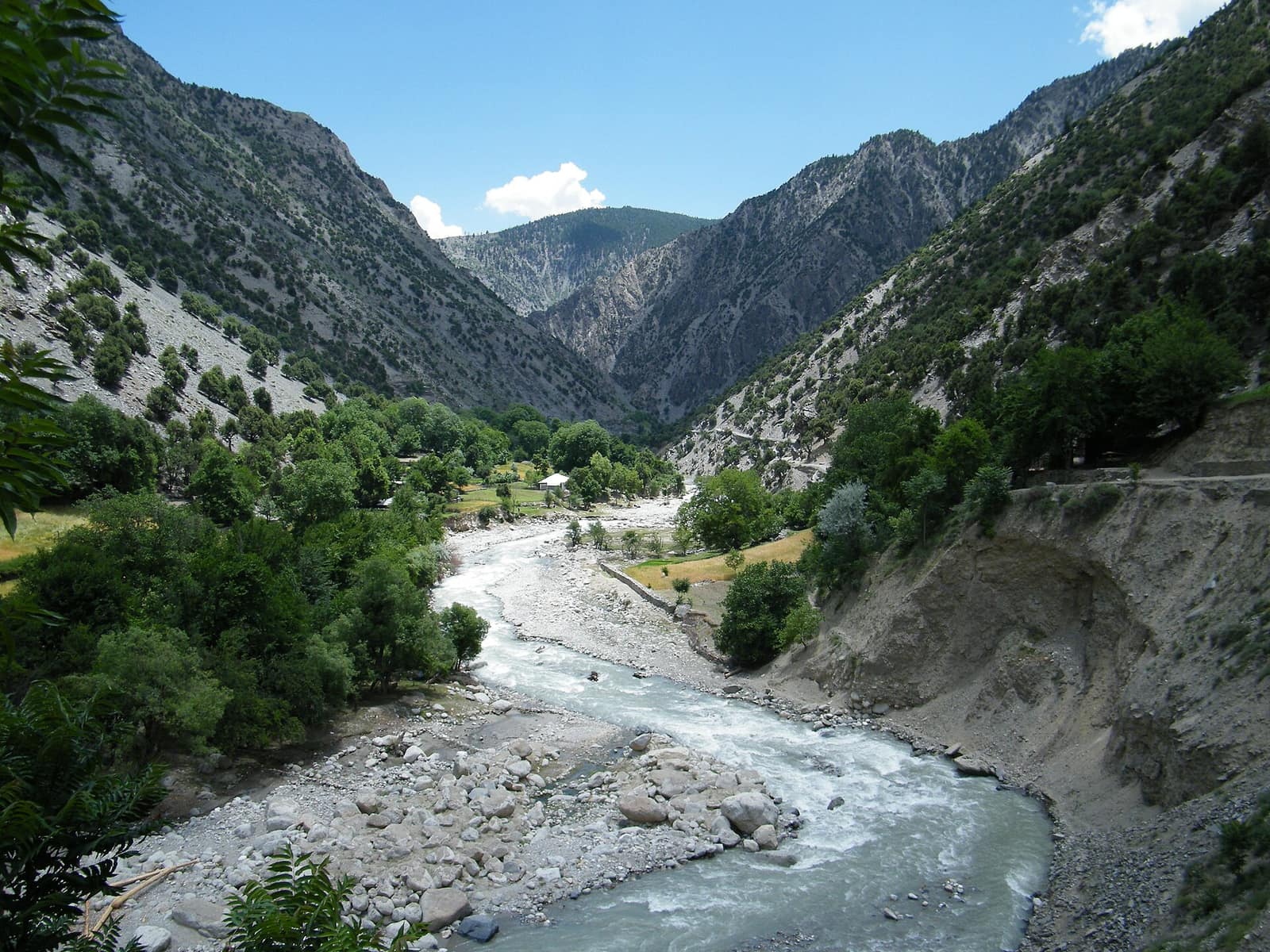 A grey-green river running over boulders through the Rumbur valley in Chitral, trees along its banks and steep bare mountains closing in behind