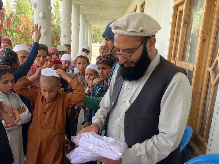 Sadiq Amiri, director of operations in Nuristan, in a white pakol, glasses and grey waistcoat, handing out new white clothes to Nuristani children on Eid, a boy beside him trying on his new cap