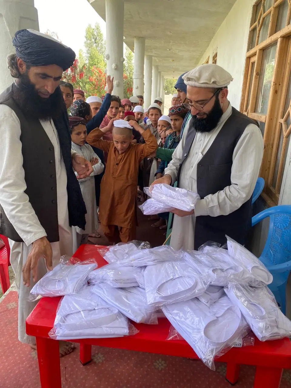Sadiq Amiri, on the right in a white pakol and glasses, and a colleague in a black turban hand out new white clothes in plastic bags from a red table to a crowd of Nuristani boys on a veranda on Eid, one boy trying on his new white cap